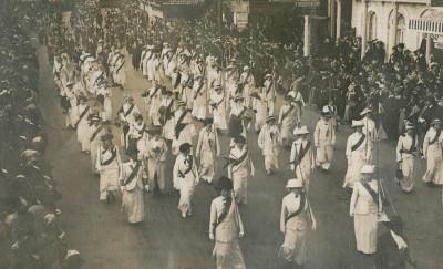 A black and white photo of a number of women marching in the street, wearing matching white outfits and dark sashes while crowds look on
