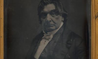 a somewhat blurry black-and-white image of a white man wearing a jacket and bowtie in a frame.