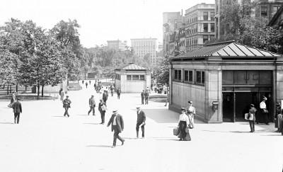 Black and white photograph depicting a subway entrance with many well-dressed men and women walking past. The photo also shows two young boys selling newspapers to the passing crowd